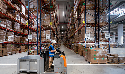 Man driving on an electric pallet truck through a large warehouse filled with inventory.